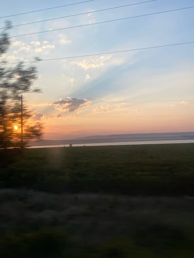 The image depicts a sunset scene over a rural landscape, likely on the outskirts of Balabanu, Moldova. In the foreground, a blurry tree branch with its leaves is visible on the left, with the sun peeking through it. The sun itself is a bright orb, casting a warm glow. Power lines are strung across the upper part of the frame, spanning the sky. The sky transitions from a pale blue at the top to shades of orange and pink near the horizon, with scattered clouds. Below the sky, a body of water is visible, possibly a river or lake. Beyond the water, a flat, dark green field stretches out, possibly a cultivated crop. In the far distance, a hazy, layered landscape of hills or mountains is visible. The overall impression is one of peaceful, natural beauty at the end of the day.