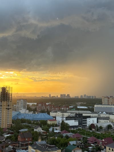 The image depicts an aerial view of a city under a dramatic sky. The sky is filled with dark, heavy clouds, particularly on the left side, with a bright, golden-yellow light breaking through from the horizon on the left. This suggests either a sunrise or sunset, with the sun's rays illuminating the clouds and casting a warm glow over a portion of the cityscape.

In the foreground, a skeletal structure of a tall building under construction is visible on the left. Below and to the right of this, the city unfolds with a mix of buildings. There are several multi-story residential buildings, some with red brick facades and others with lighter colored exteriors. A large building with a distinctive blue, domed roof is also prominent. Further into the city, more urban development can be seen, with a cluster of taller buildings in the distance, some appearing to be modern high-rises.

Interspersed with the urban structures are areas of dense green trees and vegetation, suggesting parks or forested areas within or bordering the city. In the lower right of the image, a denser residential area with smaller houses and rooftops is visible.

The overall impression is one of an active urban environment at the cusp of changing weather conditions, with the dramatic sky creating a strong visual contrast with the organized structures of the city. There are no visible people or actions occurring in the image. The location is identified as Shirkovo, Russia.