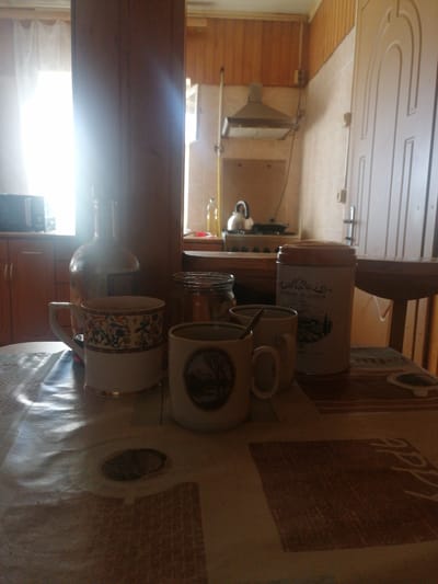 The image depicts a still life in a domestic setting, likely a kitchen or dining area in Dubrovka, Russia. The scene is arranged on a table covered with a patterned tablecloth, upon which sit several objects. In the foreground, there are three ceramic mugs, one with a decorative floral pattern and a gold rim, another with a scenic illustration, and a third plain white mug. To the right of the mugs is a cylindrical tin can with a wooden lid, featuring text that reads "HOMEMADE TRADITION" and a landscape illustration. Behind the mugs and tin can, a large glass bottle, possibly containing a beverage, stands prominently. A glass jar, partially filled, is also visible.
The background reveals a kitchen environment. A microwave is visible on a cabinet to the left. Further back, a stovetop with a kettle and a range hood are discernible, suggesting recent or ongoing food preparation. The walls are paneled with light-colored wood at the top and tiled with a neutral tone below. A door with carved detailing is on the far right. Natural light streams in from a window on the left, casting a strong glare and creating shadows, implying it is daytime. The overall atmosphere is subdued, with a focus on the collection of items on the table, giving a sense of a paused moment, perhaps before or after a meal or a gathering. The lighting and domestic setting evoke a feeling of everyday life and perhaps a touch of rustic charm.