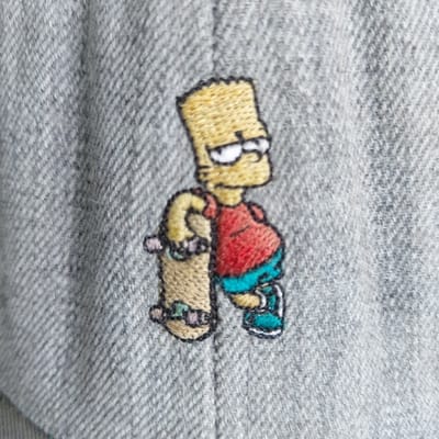 A close-up photograph displays an embroidered depiction of the animated character Bart Simpson on a grey twill fabric. Bart Simpson is rendered with yellow spiky hair, white eyes with black pupils, a red sleeveless shirt, blue shorts, and blue shoes with white soles. He is shown standing and holding a light brown skateboard with purple wheels and light blue trucks vertically against his left side with his left hand. The embroidery uses black outlines to define the character's features and form. The background consists solely of the grey fabric, which exhibits a diagonal weave pattern. The depicted object is not situated within a visually identifiable scene indicating its specific location in Gimpo-si, South Korea.