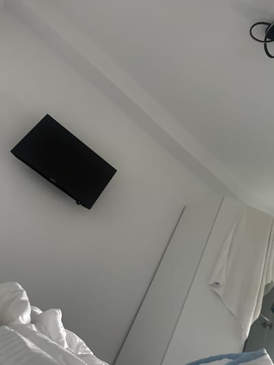 The image depicts an interior scene, likely a bedroom, angled to emphasize a wall-mounted black television. The television is positioned on a white wall, with a dark border and a small red indicator light visible at the bottom. To the right of the television, a portion of a white closet or wardrobe is visible, with a folded white towel or garment hanging from it. In the lower left corner, white bedding, possibly a duvet and pillows, is partially visible. The ceiling above is white and features angled sections. A black decorative light fixture is visible in the upper right corner. The scene appears to be clean and well-lit, with natural light casting shadows. The location is identified as Stațiunea Zoologică Marină Agigea, Romania.