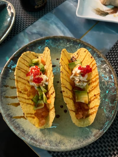 The image presents a close-up, high-angle view of two meticulously arranged appetizer-sized dishes, resembling small tacos, served on a round, artistic ceramic plate. The setting appears to be indoors, likely a restaurant, with a dark, woven placemat partially visible underneath.

Each dish features a crisp, golden-yellow shell, similar to a corn tortilla or a thin cracker, folded into a U-shape. The fillings are vibrantly colorful and layered: a central dollop of creamy white sauce or whipped topping is generously garnished with bright red roe or caviar. On one side of the cream, there are neatly diced chunks of green avocado, and on the other, a mound of finely diced, reddish-orange ingredient, which could be raw fish like tuna or salmon, or a fresh tomato salsa. A dark, glossy drizzle, possibly a balsamic glaze or a similar reduction, is artfully applied across the plate and the shells, adding visual depth and complementing the dish.

The ceramic plate itself is a prominent feature, showcasing a unique, textured design with swirling patterns and a glossy finish that reflects light, presenting a blend of blue, green, and grey tones. The lighting is atmospheric, likely artificial, creating a sophisticated ambiance. In the upper-left corner, a portion of a white rectangular dish, possibly for sushi or another accompaniment, is partially visible. Another small, dark dish is glimpsed in the lower-left. The scene captures an exquisite food presentation, emphasizing freshness and gourmet styling. No text is visible in the image. The provided context states the location is Basiglio, Italy.