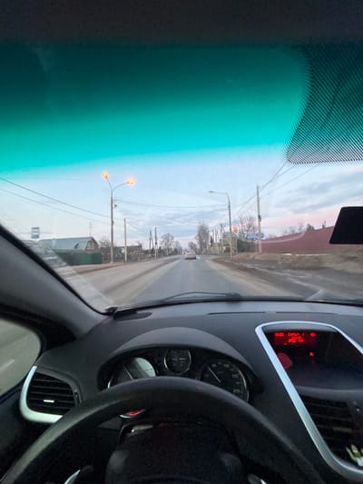 A first-person perspective from the driver's seat of a vehicle in Yasenevo, Russia, depicts a street scene at dusk or dawn. The vehicle's black steering wheel and dashboard are prominent in the foreground. A speedometer registers approximately 5-10 km/h, with a maximum reading of 210 km/h. The central console display shows "13:00", "INPUT 1", "10°C", and "25.0 KM". The upper portion of the windshield exhibits a green tint.

The two-lane asphalt road ahead is wet, with visible dirt and slush accumulating on the shoulders. Illuminated streetlights are positioned on utility poles along both sides of the road. On the left, a bus stop sign is discernible, followed by a residential building with a distinctive green roof and bare trees. On the right, additional utility poles, bare trees, residential structures, and a prolonged reddish-brown fence or wall are observed. A white sedan is visible in the distance, proceeding away from the viewer in the right lane. The sky is overcast, displaying light blue and grey tones with faint hints of pink near the horizon.