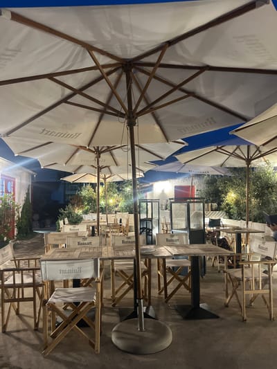 The image displays an outdoor restaurant terrace in Caminha, Portugal, at night. At least seven large, white, octagonal patio umbrellas with wooden frames are open, providing overhead coverage. Many of these umbrellas prominently feature the inverted "Ruinart" logo on their undersides. Approximately twenty light-colored wooden tables, some with dark metal bases, are arranged across the paved patio area. Numerous light-colored wooden chairs, predominantly in a director's chair style with white fabric seats and backs, accompany these tables. The white fabric chair backs consistently display the "Ruinart" logo.
The scene is illuminated by artificial ambient lighting, casting a warm glow. The ground is a uniform light-colored paving. In the background, dense green foliage is visible, interspersed with small artificial lights, indicating landscaped elements. To the far left, a light-colored building wall with a distinct red-framed doorway or window is present. In the right mid-ground, a metal and glass partition or similar structure can be observed. One individual is partially visible on the far right, seated at a table, holding what appears to be a dark menu. No other individuals are clearly identifiable. The setting appears prepared for dining but is largely unoccupied.