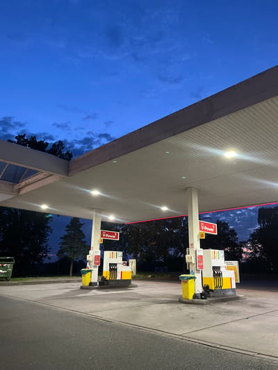 This is a low-angle, full shot of a Shell gas station at dusk in Garbsen, Germany. The sky is a deep blue with scattered, wispy clouds. The gas station canopy is prominent, lit from within by fluorescent lights. Underneath the canopy, two sets of gas pumps are visible. The pumps are white and yellow, with red "V-Power" branding. One set of pumps has three nozzles, while the other has two. Yellow trash cans are situated next to the pumps. In the background, dark silhouettes of trees line the horizon. A green dumpster is visible to the far left of the frame. The ground is paved concrete, with a grey asphalt road in the foreground. The scene is devoid of people.