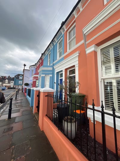The image depicts a row of colorful terraced houses in Eastbourne, United Kingdom. The houses are painted in a variety of pastel shades, including orange, blue, red, and purple. The facades feature white window frames and architectural details. Black wrought iron fences line the front of the properties.
A narrow street runs alongside the houses, with black bollards placed along the pavement. The street appears wet, possibly from recent rain. The sky is overcast with dark, heavy clouds.
Visible objects include: windows with white shutters, potted plants, refuse containers, utility poles, electrical wires, and parked vehicles in the distance. No people are visible in the image. The overall impression is of a residential street in a seaside town under a cloudy sky.