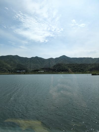 This horizontally oriented landscape image captures a serene natural scene, likely during the daytime, possibly late morning or afternoon, indicated by the bright sky and shimmering water. The view is divided into three main sections: a body of water in the foreground, a mountain range in the midground, and a sky above.
The right half of the image is dominated by a calm, deep greenish-blue body of water, which could be a lake or a wide river. Its surface is covered with gentle ripples, and numerous small, bright glints of sunlight sparkle across it, suggesting a sunny day.
Beyond the water, a range of undulating mountains stretches across the central part of the image. These mountains are densely covered in vibrant green trees and foliage, indicating a lush, forested environment. At the base of the mountains, near the waterline, several small, light-colored structures, possibly buildings, are visible, suggesting a small settlement or infrastructure. A tall, slender structure, resembling an electricity pylon, is faintly discernible on a mountain ridge in the distance.
The sky, occupying the left half of the image, is a light, clear blue, adorned with scattered white clouds. Some clouds are thin and wispy, while others appear more diffused and irregular, adding texture to the expansive sky.
The image conveys a sense of tranquility and natural beauty. No specific activity or event is taking place, and no visible text is present. The provided context indicates the location is Yangsan-si, South Korea.