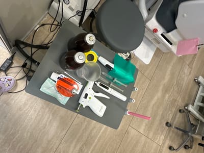 The image depicts a medical or dental setting, likely a clinic or examination room. A gray cart is laden with various medical instruments and supplies, including syringes, a dental curing light, possibly dental anesthetic capsules, and what appears to be a stapler-like device. Other items on the cart include a small plastic cup, disposable surgical instruments in a sterile package, and what could be a tray or dish with cotton swabs. A medical chair is visible, equipped with a pink surgical drape, alongside other medical equipment components. There are multiple power cords and cables running from the cart, suggesting connectivity to devices out of frame. A chair with wheels is on the left. The presence of Russian text on one of the packages suggests the location is in Russia. There is no evidence in the image to indicate the city of Kraskovo, Russia.