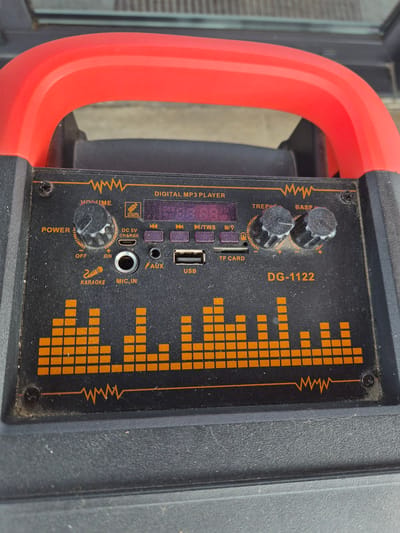 This is a close-up, eye-level, outdoor shot of a portable speaker with a bright orange handle. The speaker is black and rectangular, with a prominent equalizer display made of orange squares on the right side of its control panel. The control panel also features several knobs, buttons, and input ports.
Two knobs are labeled "TREBLE" and "BASS." Below these, a small digital screen is visible, along with buttons labeled "MODE," "M/TWS," and playback control symbols. Further down, there are ports for "TF CARD," "USB," and "AUX." Below the "USB" port, there is a "DC 5V CHARGE" input, a microphone input labeled "MIC.IN," and a switch labeled "POWER" with "OFF," "ON," and a "+" and "-" indicator. A button labeled "KARAOKE" is also present.
The speaker appears to be dusty, suggesting it might be used outdoors or has been stored for some time. The bright orange handle curves around the left side of the speaker. The background is blurred, but appears to be a gray, textured surface, possibly a wall or pavement. There are no people or animals visible in the image. The overall impression is that of a functional, though somewhat weathered, piece of audio equipment.