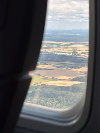 The image shows an aerial view from an airplane window of the landscape surrounding Tremblay-en-France, France. The foreground is dominated by the dark interior of the airplane, partially obscuring the view. The landscape below is a patchwork of agricultural fields, some appearing to be harvested with golden stubble, while others are green. Tree lines and patches of forest are visible, creating a textured terrain. In the distance, a town or urban area can be seen, with buildings and infrastructure. The sky is partly cloudy, with soft daylight illuminating the scene. The photograph captures a typical view of a rural and semi-urban environment from an elevated perspective.