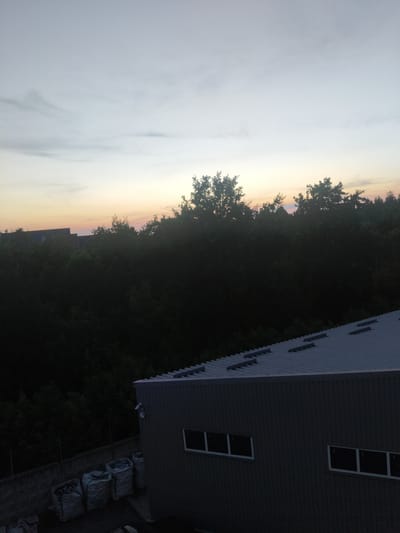 The image shows a dark, overcast sky with a hint of orange and pink near the horizon, indicating either dawn or dusk. Below the sky is a dense line of dark green trees. In the foreground, a portion of a corrugated metal roof of a building is visible, along with the side of the building which is grey and features several rectangular windows. To the left of the building, there are several large, light-colored bags, possibly filled with materials, stacked near a concrete wall. Security cameras are mounted on the side of the building. The overall scene appears to be an industrial or storage area on the outskirts of the city of Sviatopetrivske, Ukraine.