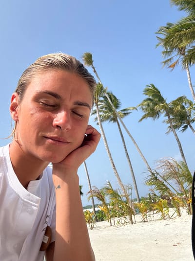 The image depicts a close-up of an individual resting outdoors on a sunny day in Magoodhoo, Maldives. The subject is a person with light-colored, slicked-back hair, appearing wet, and closed eyes. A septum piercing is visible. Their right hand is positioned beneath their chin, supporting their head. A small, dark tattoo is present on their left forearm/wrist. They are wearing a white collared shirt with visible buttons and a brown, oval-shaped tag or badge attached to the right side of the shirt. The background features a clear blue sky and numerous tall palm trees with green fronds, some appearing wind-swept or leaning. A stretch of white sand beach is visible, along with smaller green vegetation and distant, indistinct structures suggesting a resort or settlement environment.