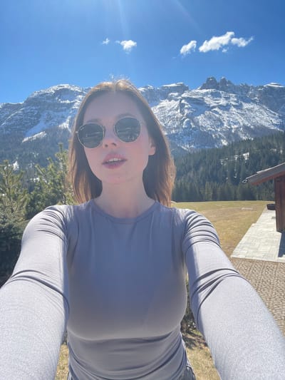 A selfie of a woman wearing sunglasses in Sappada, Italy. The woman is wearing a gray long-sleeved shirt. Snow-covered mountains are visible in the background, along with trees and a blue sky with scattered clouds. Part of a building is visible on the right side of the frame.