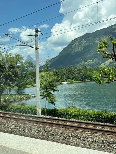 The image depicts a serene lakeside scene in Zell am See, Austria. In the foreground, a set of train tracks runs horizontally across the frame, bordered by gravel and a light-colored concrete structure. Beyond the tracks, a lush green hedge lines the shore of a calm, turquoise lake. Several people are visible on the grassy bank by the water, some sunbathing and others sitting.

The middle ground is dominated by the expansive lake, reflecting the clear blue sky dotted with white, puffy clouds. Trees line the far side of the lake, creating a verdant backdrop. In the distance, a majestic mountain range rises, its peaks partially covered in snow, indicative of the alpine surroundings. A single sailboat is visible on the lake, adding a touch of life to the tranquil scenery.

A tall, grey utility pole with electrical wires stretches vertically up the left side of the image, partially obscuring the view of the mountains. The overall atmosphere is one of peace and natural beauty, capturing a typical summer day in the Austrian Alps.