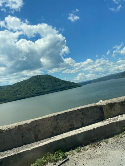 The image presents a daytime landscape capturing a wide body of water, identified as the Danube River, within the geographical vicinity of Drobeta-Turnu Severin, Romania. The upper portion of the frame is dominated by a bright blue sky partially filled with numerous white cumulus clouds of various sizes and formations.
In the immediate foreground, a weathered concrete barrier, appearing as a multi-tiered guardrail or embankment, extends horizontally across the lower part of the image. This structure exhibits a coarse, granular texture and is grey in coloration. Below the concrete barrier, a narrow strip of ground composed of dirt and gravel is visible, supporting sparse patches of green vegetation and some minor debris.
Beyond the concrete barrier, a substantial expanse of water occupies the middle ground. The water surface displays a muted greenish-grey tone and appears relatively calm. To the left, a large, steep landmass, completely covered in dense, dark green deciduous forest, rises directly from the water's edge. On the right side of the water body, several receding, forested hills or mountains are visible in the distance, extending towards the horizon under the cloud-strewn sky.
No human subjects, animals, or man-made structures other than the concrete barrier and distant hints of settlement on the far shore are discernible within the frame. The lighting suggests direct sunlight from a clear sky.