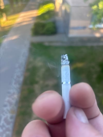 A close-up, low-angle shot shows a lit cigarette being held between the thumb and index finger of a person, with the other fingers blurred in the foreground. The cigarette is white with a silver band, and a small amount of ash and glowing ember is visible at the tip. Thin wisps of smoke curl upwards from the cigarette. The background is out of focus, showing a blurred view of an outdoor environment with patches of green foliage and a paved walkway, suggesting a park or residential area in Pereiaslav, Ukraine. Sunlight illuminates the scene, creating bright highlights on the walkway and foliage.