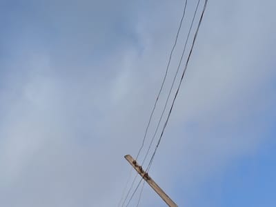 The scene, located in Adikpo, Nigeria, captures an upward view of the sky. The majority of the frame is occupied by a light blue sky with diffuse white clouds scattered throughout. In the lower-right quadrant, a weathered wooden utility pole is visible, extending diagonally upwards and towards the left. Three dark-colored electrical or communication wires are strung parallel to each other, attached to the visible portion of the pole, and traverse the sky from the lower-left to the upper-left section of the image. There are no people, explicit actions, or additional significant objects visible within the frame.