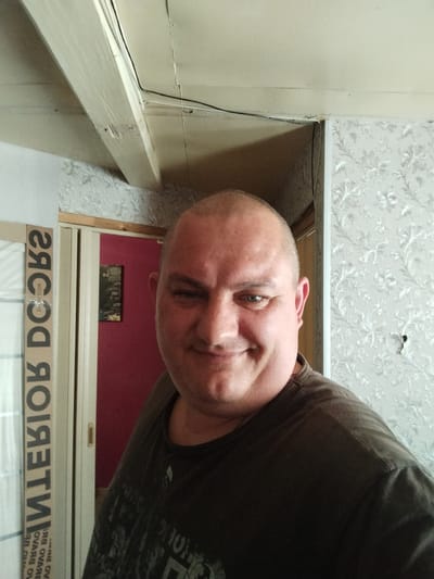 A man with a shaved head and light blue eyes is taking a selfie indoors. He is wearing a dark t-shirt. Behind him, a door is visible, painted a deep red, with a framed picture on it. To his left, a white door frame with the word "DOORS" printed vertically in black letters is visible. The walls are covered in a floral patterned wallpaper. The ceiling appears to be made of wood beams and panels. The scene is likely within a residential building.