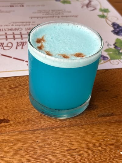 A close-up, eye-level, medium shot captures a tall, clear glass filled with a bright blue beverage, topped with a frothy white foam. The foam is sprinkled with a light dusting of cinnamon in a splotchy pattern. The glass is positioned on a wooden table with a prominent wood grain. In the background, a pinkish-white menu is visible, featuring decorative illustrations of grapevines and grapes on the right side, and what appears to be text and vertical lines on the left. A small portion of the menu reads "del Rio" and "del CAMPESINO". The overall setting suggests a restaurant or cafe in San Vicente de Azpitia, Peru, a region known for its agricultural products and vineyards. The lighting is natural, with a slight shadow cast on the wooden surface to the left of the glass. There are no people present in the frame.