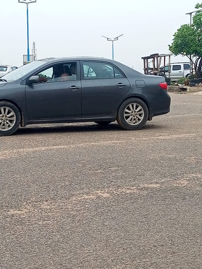 A gray sedan is parked on a coarse gray asphalt road in what appears to be Oshogbo, Nigeria, as suggested by the urban setting and the style of streetlights and roadside structures in the background. Inside the car, a person is visible in the driver's seat wearing patterned clothing. Behind the car, a white pickup truck can be seen parked near a simple wooden structure. A small tree is next to the wooden structure. Tall streetlights are also visible in the background.