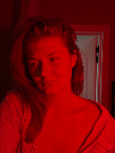 A female subject is depicted indoors within the city of Tashevka, Russia, under pervasive red light. The subject, possessing light skin and medium-length hair, is centrally positioned, with her head angled slightly to the left and gaze directed off-frame left. A faint smile is visible on her face. She wears a light-colored, textured, long-sleeved top with a wide neckline, exposing a portion of her right shoulder and collarbone. In the background, to the upper right of the subject, a white doorframe with a visible dark hinge is observed against a red-lit wall. The background to the subject's immediate left is dark and indistinct.