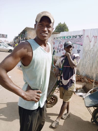 This image depicts an outdoor, brightly lit street scene in Mumbwa, Zambia. Two adult dark-skinned males are positioned in the foreground.
The man on the left stands facing forward, looking directly at the camera with a slight smile. He wears a light-colored (possibly pale blue or green) tank top, dark trousers, and a light brown baseball cap. His left hand rests on his hip, and perspiration is visible on his arm and neck.
The man on the right stands slightly behind and to the right of the first man, leaning on a pushcart. He wears a multi-colored, patterned t-shirt, light brown shorts, and a dark baseball cap. He is looking towards the right side of the frame with a slight smile, his right hand near his chin. He wears light-colored sandals.
Between the two men is a pushcart or wheelbarrow, with another partially visible on the far right. Behind the man on the right is a corrugated metal fence serving as a backdrop. Affixed to this fence is a white sign with green lettering, advertising "ANTHONY MOTHERCARE BABY'S SHOP," featuring images of baby products and contact information, including a phone number beginning with "+234". To the left, in the background, a street is visible with parked vehicles, including a white sedan, and multi-story buildings. Further in the distance, a billboard structure is discernible. The ground is unpaved and appears dusty, with some rubble near the base of the corrugated fence. Green foliage from trees is visible above the background structures. The scene suggests a commercial or market area under clear daylight.