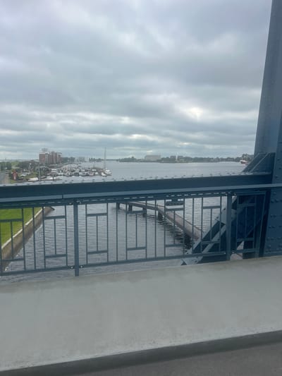 An overcast day in Wilhelmshaven, Germany, features a view from a bridge overlooking a harbor. The foreground shows the concrete edge of the bridge and a dark blue metal railing. Beyond the railing, a body of water is visible, with moored sailboats and yachts along the left bank. Several buildings, including a multi-story brick structure and smaller houses, line the waterfront. In the distance, the expansive water continues, with a large, light-colored building visible on the far shore. The sky is filled with dense, grey clouds, suggesting a potentially damp or windy day. The scene captures a typical harbor environment with a mix of maritime activity and urban development under a somber sky.