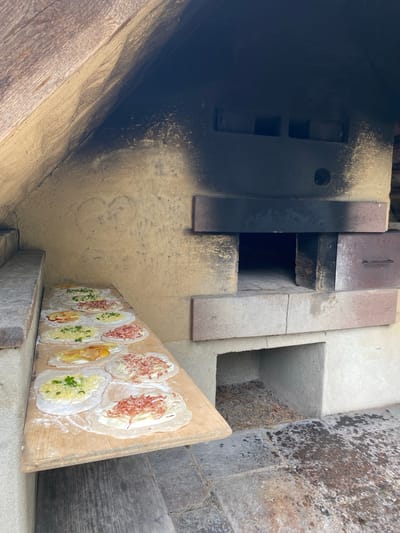 Here is a concise description of the image:

The image shows a wooden peel, likely for pizza or flatbread, with several uncooked pizzas topped with various ingredients. The peel rests on stone or concrete steps, leading to what appears to be a wood-fired oven, as evidenced by the dark soot stains around the opening. The oven is built of brick and stone and is in Gutach (Schwarzwaldbahn), Germany.