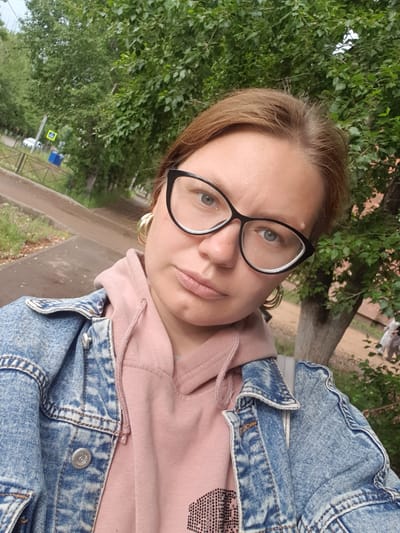A selfie of a woman in Ust-Ordynsky, Russia, taken outdoors in daylight. The woman has light brown hair, is wearing large black-rimmed glasses, and large gold hoop earrings. She is wearing a pink hooded sweatshirt under a distressed denim jacket. The background shows a sidewalk, trees with green foliage, and a street with a yellow pedestrian crossing sign and a blue trash bin. The overall setting appears to be a residential or park area.