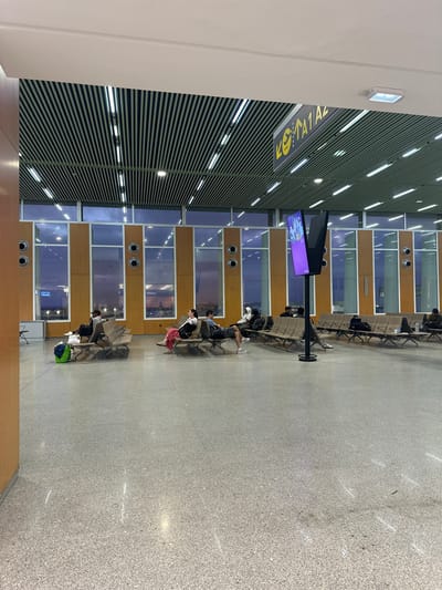 The image depicts a spacious, modern indoor waiting area, likely an airport terminal or a large transit lounge, given the context of Nouaceur, Morocco. The scene is illuminated by artificial lighting, suggesting it is evening or night.

The space features several rows of metal and upholstered benches where numerous individuals are seated, appearing to be waiting. Many have personal belongings like backpacks and bags beside them. In the foreground, a prominent black stand holds two vertical purple-hued screens, possibly displaying flight information or advertisements.

Architecturally, the area has a distinctive design. The ceiling is characterized by horizontal dark slats interspersed with long, bright strip lights. The far wall incorporates large windows, some reflecting blurred indoor lights and shapes, alternating with sections of warm-toned wooden paneling that feature circular vents. The floor is a light gray, granular surface, possibly terrazzo or polished concrete.

Notable details include:
*   A partially visible yellow sign with black text on the left side of the frame. The text appears reversed, resembling characters like "Z1A1A" or similar alphanumeric combinations, typical for gate or section identifiers in an airport.
*   Reflections in the windows and the glossy floor surface from the interior lights.
*   The overall atmosphere is quiet and organized, characteristic of a waiting area.