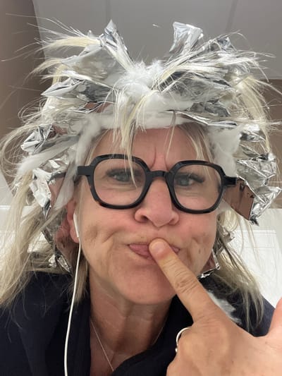 A middle-aged person wearing glasses and with aluminum foil and cotton strips in their blonde hair is shown in a close-up selfie-style photograph. The person's index finger, adorned with a ring, touches their cheek. A white earbud cord is visible near their mouth. The person wears a dark-colored top. No specific location is identifiable in the image, and there is no discernible connection to Montrond-les-Bains, France.