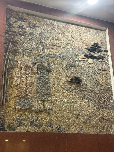 This image displays a large, rectangular pebble mosaic art piece set into a wall in Ngũ Hành Sơn Ward, Vietnam. The mural depicts a traditional Vietnamese landscape scene. On the left, stylized dark bamboo stalks with leaves extend upwards. In the center, two human figures, rendered in traditional attire using varying shades of pebbles, stand facing each other, suggesting an interaction or conversation. The figure on the left is adorned in lighter, brownish-red hues, while the figure on the right is in darker, gray-brown tones. To the right of these figures, a smaller individual rides an animal, possibly an ox or water buffalo, depicted in dark pebbles. Further right and slightly lower, two stylized houses or huts with distinctive roofs are visible. The upper right portion of the mural illustrates a sky or sunrise/sunset with radiating lines created by alternating light and dark pebbles. The entire landscape, including the ground and undulating hills, is composed of numerous small, rounded pebbles of cream, light brown, and gray hues. The mosaic is framed by dark reddish-brown polished material at the top, bottom, and right, with an overhead light source visible in the upper right corner of the overall frame.