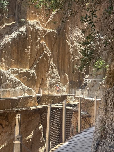 This media file depicts a scene along a narrow, cliff-side walkway constructed with metal grating and wooden planks, suspended high above a rugged, rocky terrain. The walkway features a metal safety railing with a mesh infill. Several individuals are visible on the walkway. One person, wearing a pink shirt and blue helmet, is ascending the walkway. Another person, further up, appears to be wearing blue clothing and is also on the walkway. The sun casts strong shadows, suggesting it is likely daytime, and the lighting indicates fair weather. The rocky landscape is textured with layers and crevices, with some sparse vegetation clinging to the rocks, particularly in the upper left of the frame. The overall impression is of a challenging and scenic outdoor adventure, possibly a hiking trail or a tourist attraction built into a natural canyon or gorge. The location cues suggest a place like the Caminito del Rey in Antequera, Spain, known for its dramatic natural formations and the suspended pathways. No visible text is present in the image.