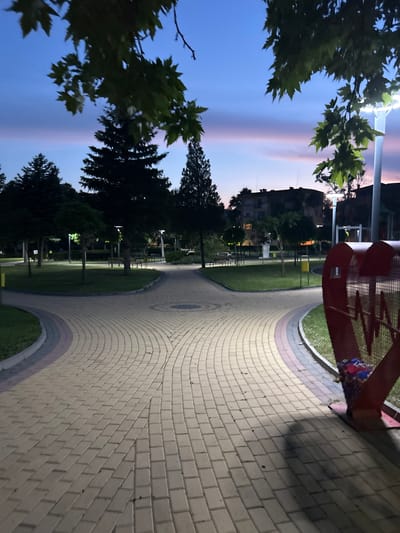 The image depicts a park in Dzhebel, Bulgaria, at twilight, characterized by a sky transitioning from deep blue overhead to soft pink and purple hues near the horizon. The park features a network of paved pathways constructed from light-colored interlocking pavers, with darker decorative strips along the edges, forming curves and intersections. Lush green lawns border these pathways, which are lined with various trees, including dark evergreen conifers and deciduous trees. Multiple modern park lights with white LED illumination are active, casting light onto the paths and surrounding vegetation, creating distinct shadows.
In the immediate foreground on the right, a large, red, metallic, heart-shaped structure is present. This structure features cutout letters, appearing to be a stylized "WM" or similar design, and functions as a collection bin, containing colorful plastic items visible in its lower section. Further into the park, several benches are visible along the pathways and on the grass. A yellow cylindrical trash receptacle is situated near a path intersection in the midground. In the more distant background, beyond the park's trees, dark silhouettes of multi-story buildings are visible against the twilight sky. No individuals are present in the scene.