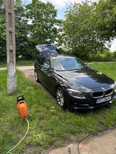 A black BMW with Bulgarian license plate "PP 2186 BT" is parked on green grass next to a gray concrete curb or small road. An orange pressure washer with attached yellow hose sits on the grass near the car. Trees are visible in the background. The location is likely Lipnik, Bulgaria, based on the license plate and user prompt.