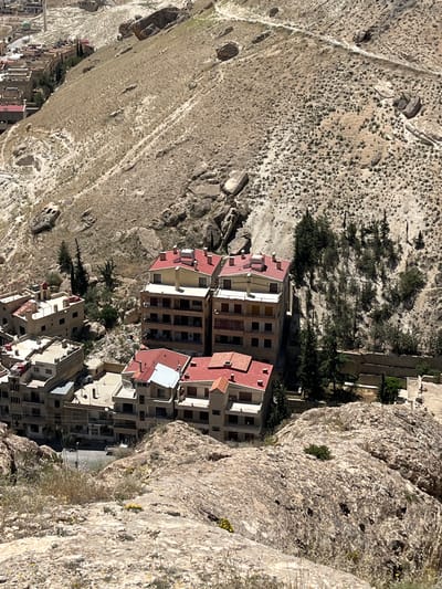 The image shows a portion of the city of Ma'loula, Syria, nestled within a mountainous, rocky terrain. The architecture consists primarily of multi-story buildings with red-tiled roofs and beige or white facades. Some buildings are directly adjacent to rock formations, while others are surrounded by sparse vegetation, including trees. The landscape surrounding the buildings is arid and rugged, with exposed rock surfaces and scattered vegetation. A road runs through the city.