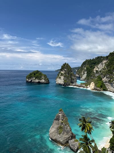 The image captures a coastal scene in Pejukutan, Indonesia, featuring a clear blue sky with scattered white clouds. The turquoise ocean dominates the lower portion, with visible variations in color indicating different depths and wave patterns. Several rocky islands of varying sizes emerge from the sea, covered in green vegetation. Palm trees line the edge of a sandy beach at the top right. The coastline consists of tall cliffs, also covered in dense greenery.