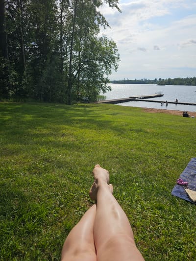 The image depicts a sunny day at a lake with a grassy area in the foreground. In the foreground, a person's legs and feet are visible, resting on the grass. Beyond the grass, a wooden pier extends into the shimmering water. Several people are present in and around the water, some swimming and others standing on the pier. Trees line the far side of the lake, with a clear sky overhead. The scene suggests a recreational area, likely a beach or swimming spot, in the city of Smålandsstenar, Sweden.