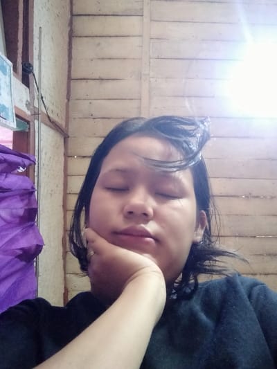 A young woman with dark hair and closed eyes rests her face on her hand. She appears calm or asleep. The background features a wall of light-colored wooden planks and a corner with purple fabric. Bright light shines from the top right corner, possibly from a window or lighting fixture. She wears a dark-colored shirt. A ring is visible on her finger.