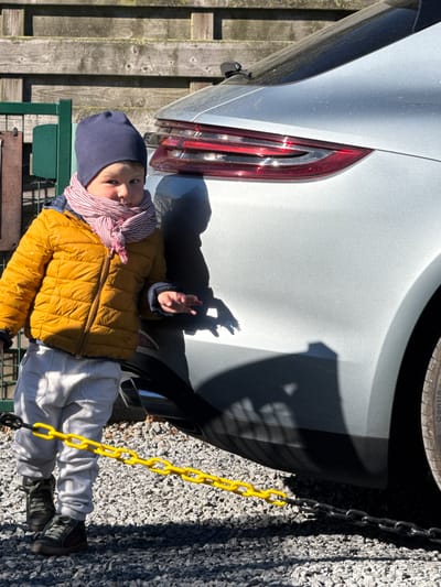 An outdoor shot depicts a young child standing on a gravel surface next to the rear left side of a silver car, with a wooden fence and a green object in the background. The image is bathed in bright sunlight, casting long, distinct shadows, suggesting early morning or late afternoon on a clear day.
The child, appearing to be a toddler or preschooler, faces the camera directly with a neutral to slightly curious expression. They are dressed warmly in a navy blue beanie, a striped red and white scarf, a puffy mustard yellow winter jacket, light grey pants, and dark sneakers with white soles. The child's right hand (viewer's left) is slightly raised, palm open.
The silver car's rear quarter panel and a section of its red taillight are prominent on the left side of the frame. Reflections of the sun are visible within the taillight. A small black sensor or camera is visible on the lower bumper. The top of a car tire, with the brand "Continental" faintly visible on its tread, is at the upper edge.
A bright yellow plastic chain runs vertically along the right side of the frame, partially connected to a black chain section at the top and bottom, which appears to be a barrier or a decorative element. The ground is covered in light-colored gravel. In the bottom left, behind the child, a part of a dark green bench or table is visible, next to a weathered wooden fence composed of vertical planks. The scene gives the impression of a casual outdoor setting, possibly a driveway or garden area in Bredene, Belgium.