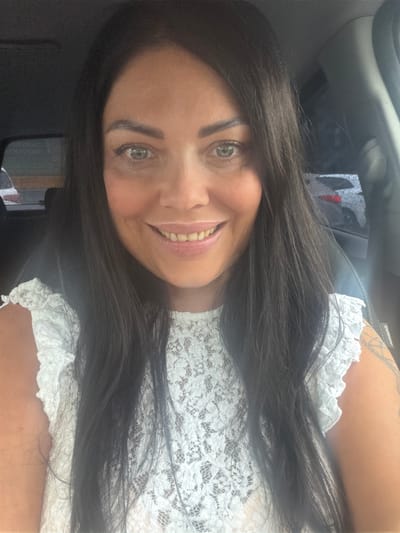 A woman with long, dark hair and light eyes is smiling at the camera. She is wearing a white lace top. The setting appears to be inside a car. There is no information to determine if the image was taken in Murmansk, Russia.