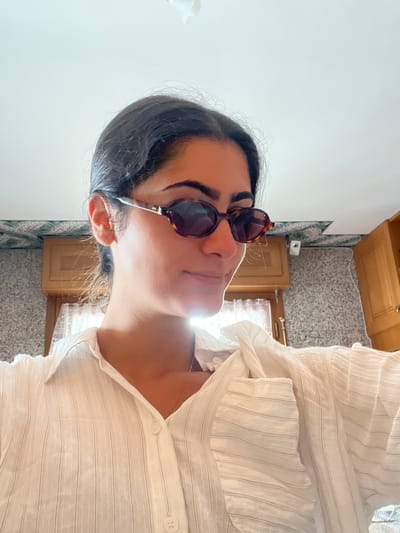 A woman with dark hair pulled back and wearing sunglasses and a white striped shirt is in an indoor setting. The background shows a kitchen area with cabinets and a tiled wall. Based on the architecture and ambiance, the location could be a residence or rental property in Èze, France.