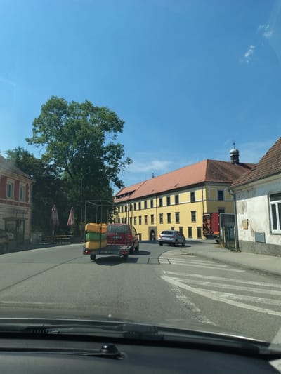 The image captures a street scene in Střelské Hoštice, Czechia, under a clear blue sky. In the foreground, the dashboard of a vehicle is visible, suggesting the photo was taken from a car. The road ahead shows a pedestrian crossing. A red car is pulling a trailer loaded with what appear to be yellow and green inflatable objects. To the left, a red and yellow building with windows and an awning is present. Further down the street, a large yellow building with a red tiled roof and numerous windows stands prominently. Several trees, including a large, leafy one, are visible throughout the scene. The environment suggests a typical European town or village setting on a sunny day.