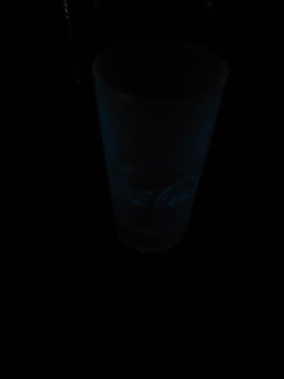 A partially visible, cylindrical object with a luminescent, blue-green glow stands out against the predominantly black background. The object's surface displays some indistinct patterns. A line of small, faint dots is visible in the lower-left corner. The image location is Oyten, Germany, though there are no indicators visible in the image itself.