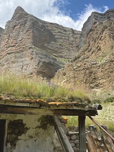 In the village of El'tiubiu, Russia, a close-up shot captures a section of a decaying, rustic dwelling. The weathered stone and plaster walls are crumbling, revealing the underlying rock construction. A dilapidated wooden roof, adorned with a thick layer of overgrown grass and small wildflowers, partially shelters the structure. The foreground is dominated by this crumbling building and its lush, green roof, while the background dramatically showcases the immense, layered rock formations of the surrounding mountainous terrain. The sky above is a mixture of blue and white clouds, suggesting a clear but potentially breezy day.