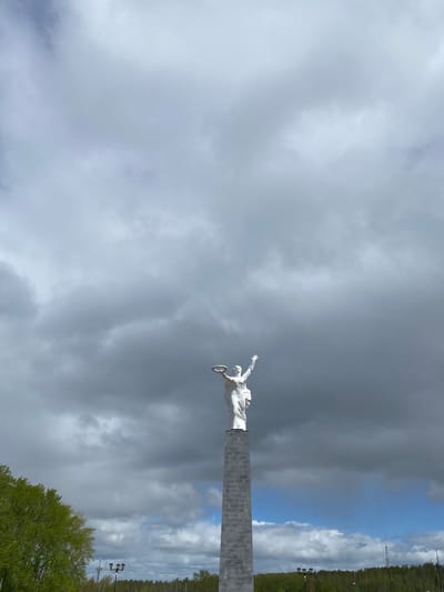 A statue of a figure holding a wreath stands atop a brick column against a cloudy sky. The column is situated near a wooded area and street lights are visible. The statue is located in Kirovgrad, Russia.