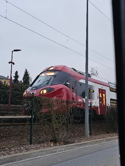 A red and black train sits beside a fenced area, with its lights on. The train car has the number 2410 displayed. The photo is taken at a slight angle and includes a gray sky and greenery in the background. A pole stands next to the train tracks, with overhead power lines running above.