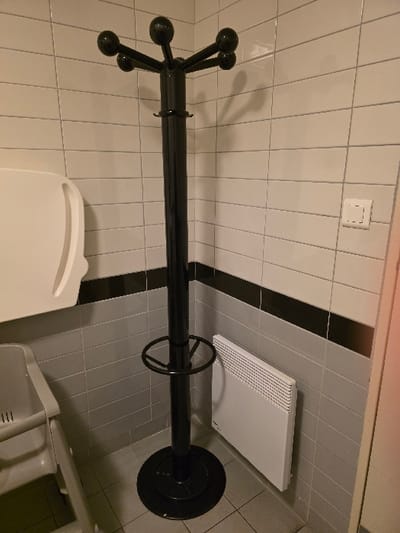 A black coat rack stands against a tiled wall, featuring a round base and multiple arms with spherical ends. Above it is a light switch. The floor is also tiled, with a white electric heater mounted on the wall. A white toilet seat is positioned in the lower portion of the frame, along with a partially visible white plastic chair.