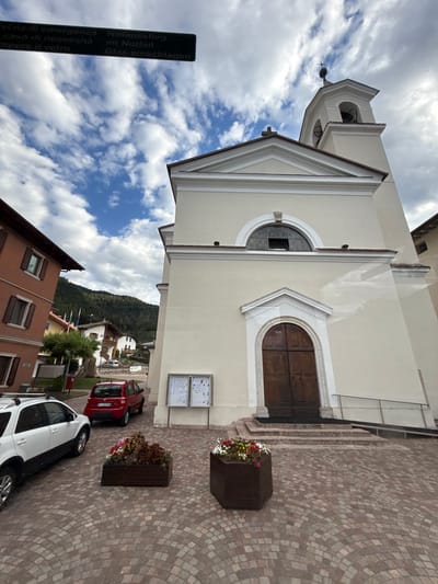 The image shows a street scene in the Italian town of Cavedago. In the foreground, there are two cars parked on a cobblestone street. The car on the left is a white SUV, and the car on the right is a red hatchback. In the middle ground, there is a church with a tall bell tower. The church is painted a light yellow color and has a large wooden door. In front of the church, there are two large planters filled with flowers. In the background, there are several buildings, including a red-colored building to the left of the church, and a backdrop of forested mountains. The sky is partly cloudy with blue patches. A sign in Italian and German is visible at the top left of the image, indicating emergency exit and emergency contact information. The scene appears to be a quiet, picturesque town square.