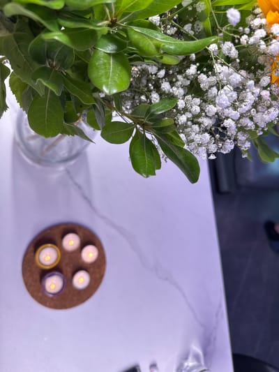 The image presents a close-up view of a tabletop still life. A bouquet of flowers, primarily white baby's breath and green leaves, sits in a clear glass vase on a white marble-patterned surface. To the right of the vase, a circular brown coaster holds five small lit candles. Two of the candles have colored holders, one yellow and one purple. The background is softly blurred, revealing a glimpse of furniture, possibly a sofa, and a dark surface. The image does not provide enough information to pinpoint the location in the city of Ariel or any other detail about the location. No people, actions, or interactions are visible in the image.